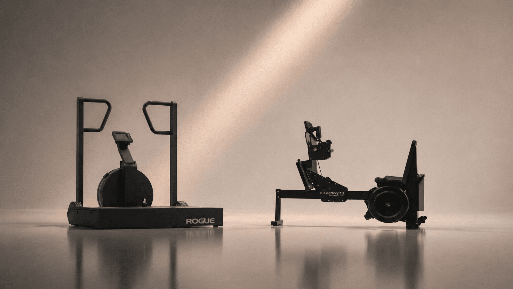 Rogue assault bike and Concept2 rower side by side under warm directional light, equipment-only register.